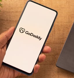 Hand holding a mobile phone showing a white screen with the GoDaddy logo