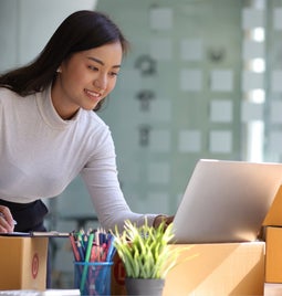 Female Entrepreneur looks at her laptop while making notes surrounded by order boxes