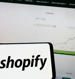 A phone with the shopify logo on it in front of a laptop screen with a graph