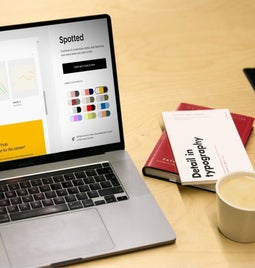 Open laptop displaying a graphic design website with design settings for color palettes, alongside a notebook, tablet, and cup of coffee on a wooden desk.