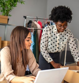 Photo of two women around a laptop and parcel boxes managing their ecommerce site