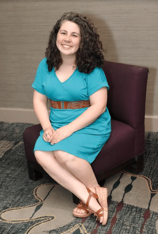 An image of Vicky Weber, wearing a blue belted dress, sitting on a purple chair.