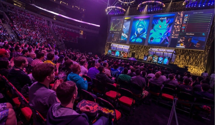 Audience watching an esports tournament with large screens displaying video game characters and player profiles.