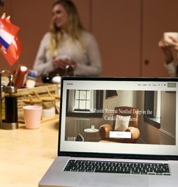 People chat in a kitchen setting with international flags, with a laptop in the foreground showing a rustic retreat in the Catskill Mountains.