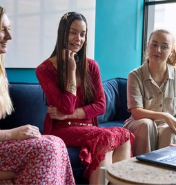 Three women in a casual business setting, engaging in conversation with a laptop on the table.