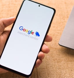 Hand holding a mobile with Google merchant center logo displayed on a white screen