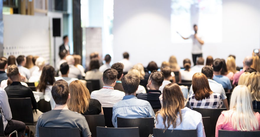 Audience of people sat in chairs listening to a speaker deliver a talk at a conference event