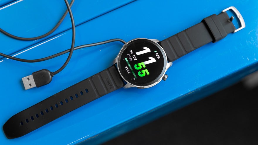 A black smartwatch with a digital display showing 11:55 is placed on a blue surface next to a USB charging cable.