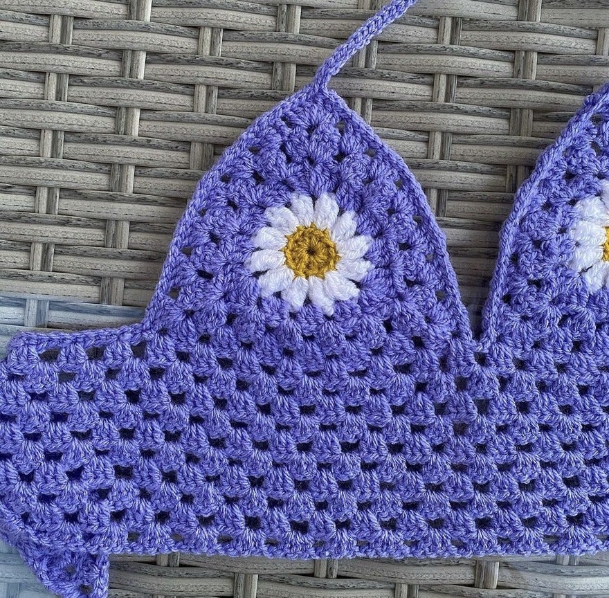 Close-up of a purple crocheted fabric with a white and yellow flower pattern, resting on a woven surface.