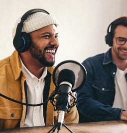 Two men sat a desk with podcast equipment in front of them and headphones on